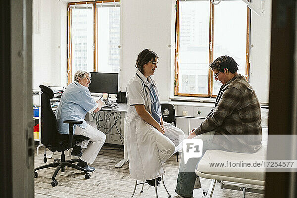 Smiling female healthcare worker talking with young male patient while senior nurse sitting at desk