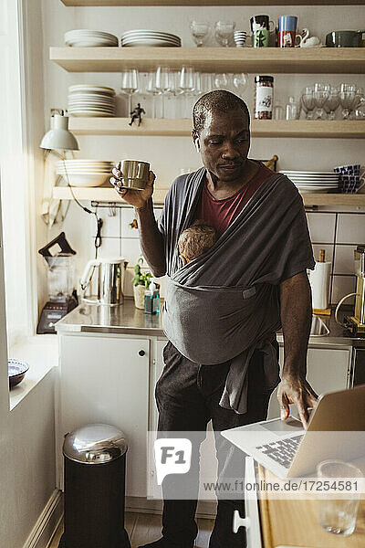 Geschäftsmann mit Sohn in Babytrage mit Laptop  während er eine Tasse in der Küche hält