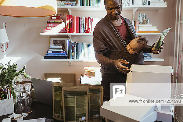 Männlicher Unternehmer  der Kisten untersucht  während er einen kleinen Jungen in der Trage im Büro zu Hause trägt