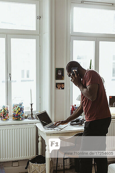 Seitenansicht eines Geschäftsmannes  der mit einem Smartphone spricht  während er einen Laptop zu Hause benutzt