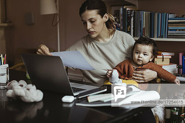 Weibliche Fachkraft liest Dokument  während sie mit männlichem Kleinkind zu Hause sitzt