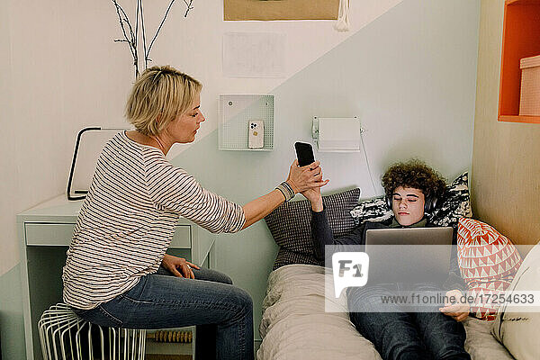 Boy giving smart phone to mother while lying with laptop on bed t home
