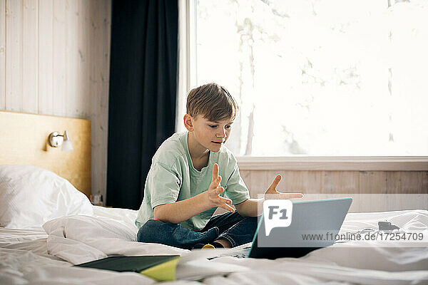 Junge nimmt an Videoanruf teil und gestikuliert während des Hausunterrichts im Schlafzimmer