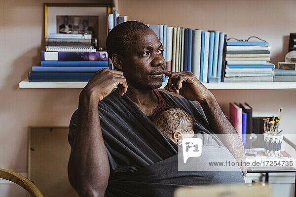 Father contemplating while sitting with son in baby carrier at home