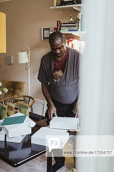 Male entrepreneur with son in carrier working on boxes at home office