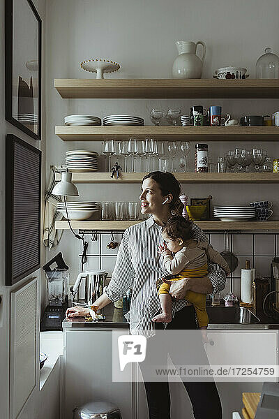 Lächelnde Mutter schaut weg  während sie ein männliches Kleinkind in der Küche trägt