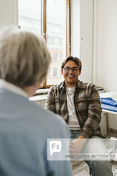 Lächelnder männlicher Patient  der einen älteren weiblichen medizinischen Experten während einer Konsultation in der Klinik ansieht