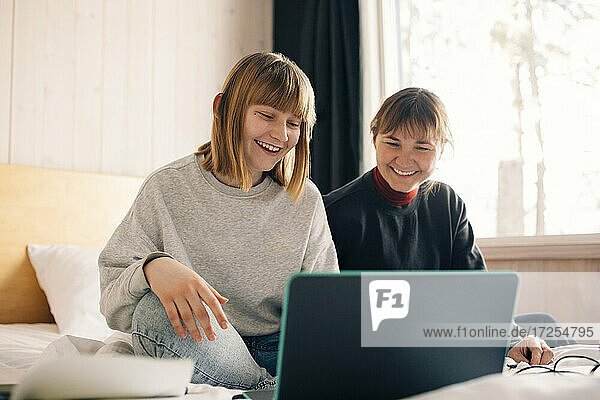 Lächelnde Mutter und Tochter sitzen mit Laptop während Homeschooling im Schlafzimmer
