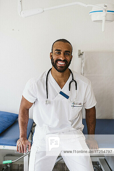 Porträt eines lächelnden medizinischen Experten  der sich auf einen Untersuchungstisch in einer medizinischen Klinik stützt