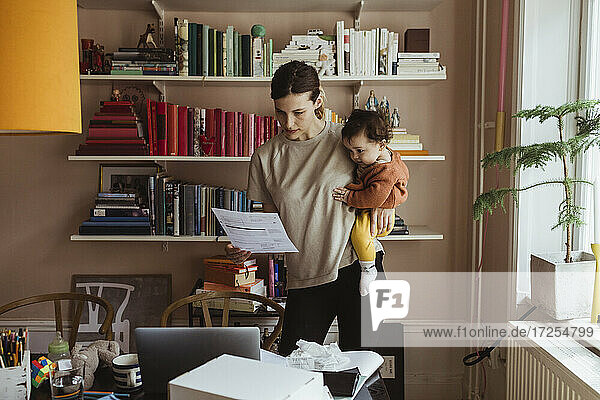 Weiblicher Profi  der ein Dokument liest  während er seinen Sohn im Büro zu Hause trägt