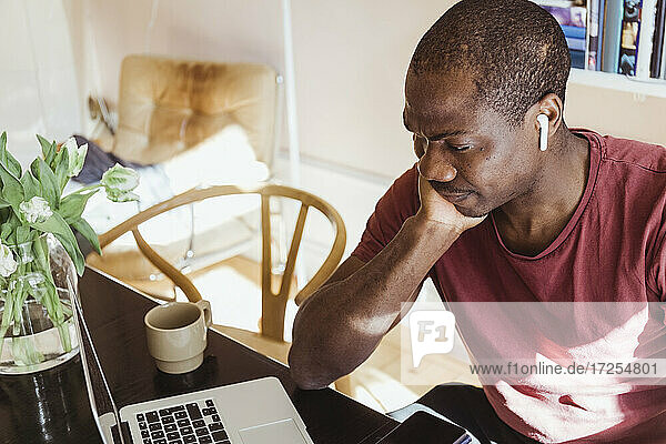 Besorgter männlicher Unternehmer mit Hand am Kinn  der auf seinen Laptop schaut  während er von seinem Heimbüro aus arbeitet