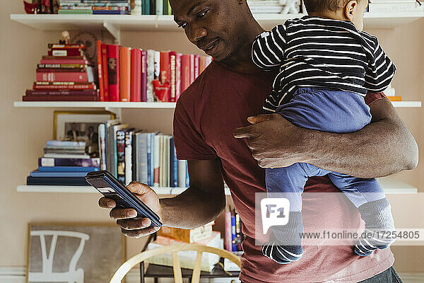 Vater verwendet Smartphone  während er einen kleinen Jungen zu Hause trägt