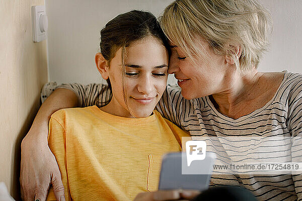 Glückliche Mutter umarmt Tochter mit Mobiltelefon im Schlafzimmer