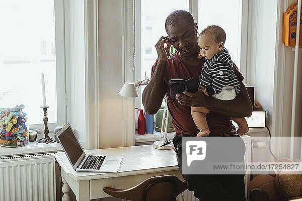 Männlicher Unternehmer  der ein Smartphone benutzt  während er einen kleinen Jungen zu Hause im Büro trägt