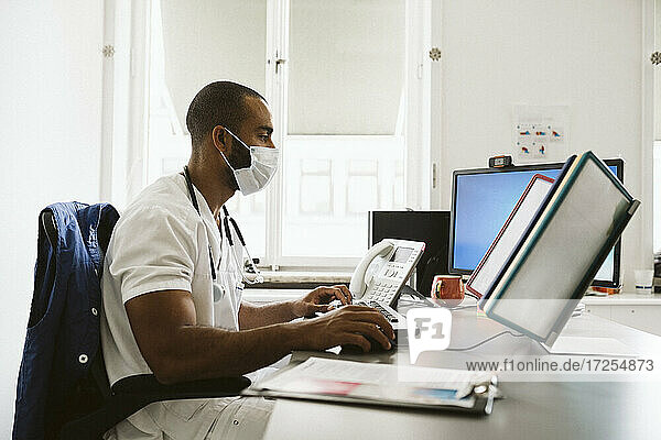 Männlicher Arzt  der einen Computer benutzt  während er am Schreibtisch sitzt  während COVID-19