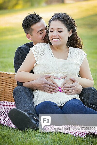 Pregnant hispanic couple making heart shape with hands on belly in the park outdoors
