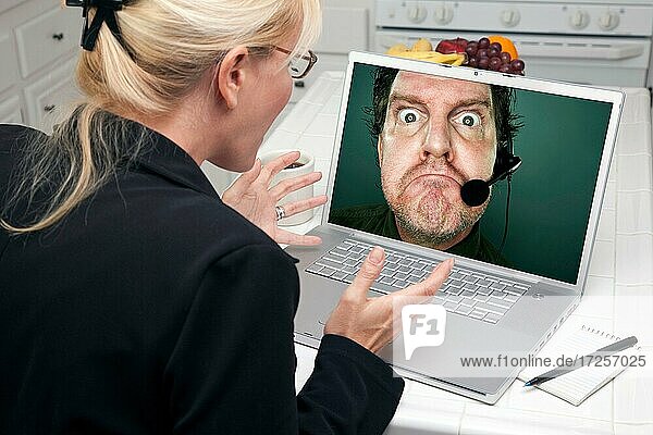 Schockierte Frau in der Küche mit Laptop mit mürrischen Kundenbetreuer auf dem Bildschirm
