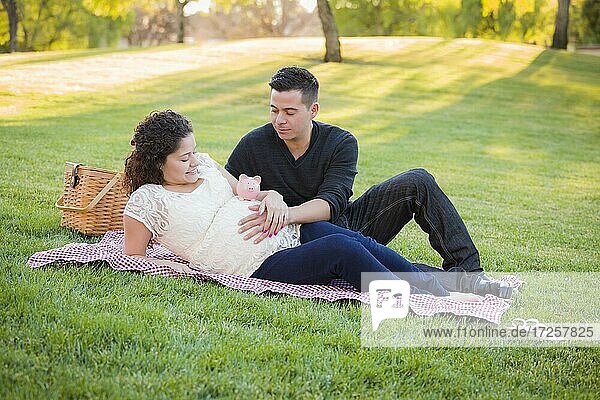 Contemplative pregnant hispanic couple with piggy bank on mother's belly in the park