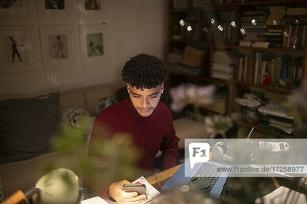 Young man with smart phone working from home at laptop in home office
