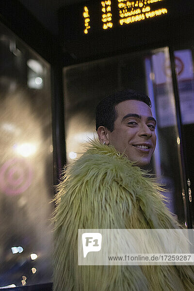 Portrait happy eccentric young man in feather coat at city bus stop