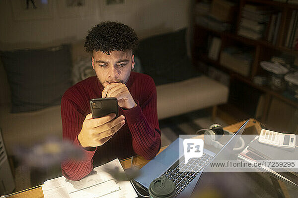 Young man with smart phone working from home at laptop
