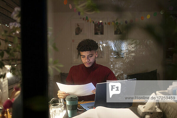 Konzentrierter junger Mann mit Papierkram  der nachts von zu Hause aus am Laptop arbeitet
