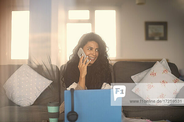 Lächelnde Frau  die von zu Hause aus arbeitet und mit einem Smartphone am Laptop spricht