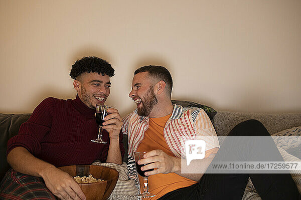 Happy gay male couple drinking red wine and eating popcorn at home