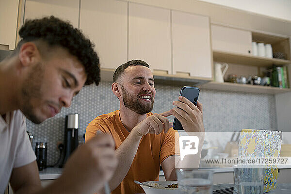 Happy gay male couple using smart phone and eating in kitchen