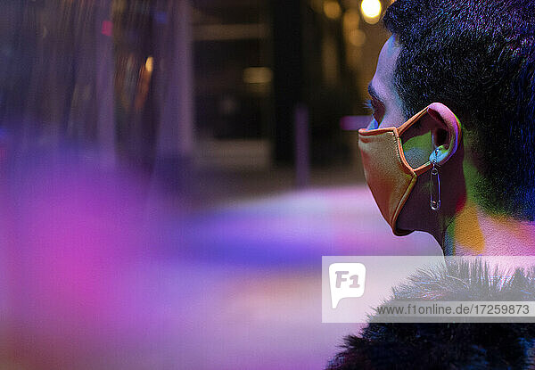 Stilvoller junger Mann mit Sicherheitsnadel-Ohrring trägt Maske unter Neon