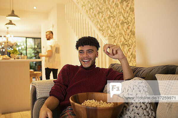Glücklicher junger Mann mit Popcorn vor dem Fernseher auf dem Wohnzimmersofa