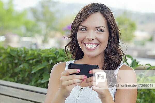Attraktive lächelnde junge erwachsene Frau schreibt auf Handy im Freien auf einer Bank