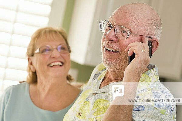 Happy senior adult husband on cell phone with wife behind in kitchen