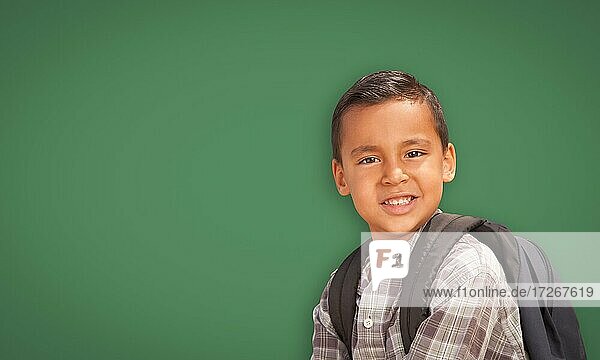 Netter hispanischer Junge mit Rucksack vor leerer Kreidetafel