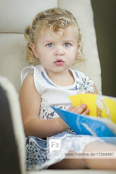 Adorable blonde haired blue eyed little girl reading her book in the chair