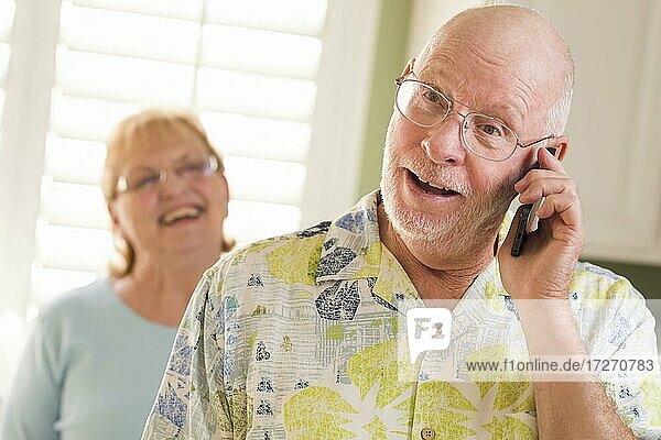 Happy senior adult husband on cell phone with wife behind in kitchen