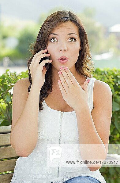 Schockierte junge adultes Weibchen  die im Freien auf einer Bank mit einem Handy telefoniert