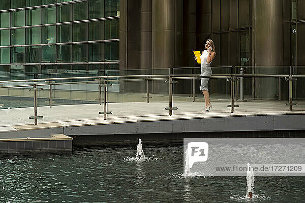 Geschäftsfrau  die über ihr Smartphone spricht  während sie auf dem Fußweg bei einem Brunnen in der Stadt spazieren geht