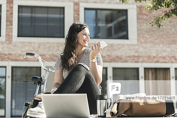 Lächelnde Frau  die über ein Smartphone spricht  während sie an einem Gebäude in der Stadt sitzt