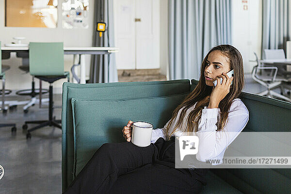 Geschäftsfrau  die mit dem Handy telefoniert  während sie auf dem Sofa im Büro Kaffee trinkt