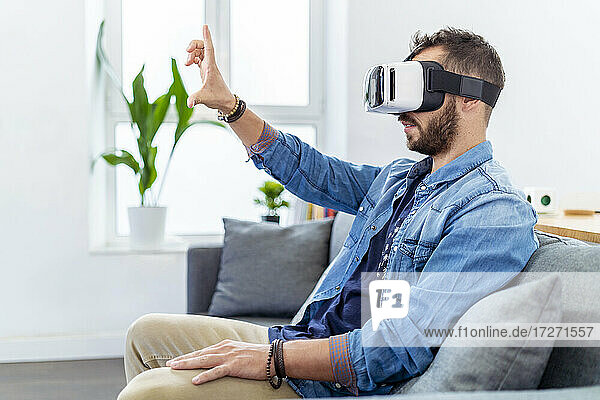 Mittlerer erwachsener Mann  der einen Virtual-Reality-Simulator trägt und auf dem Sofa sitzt