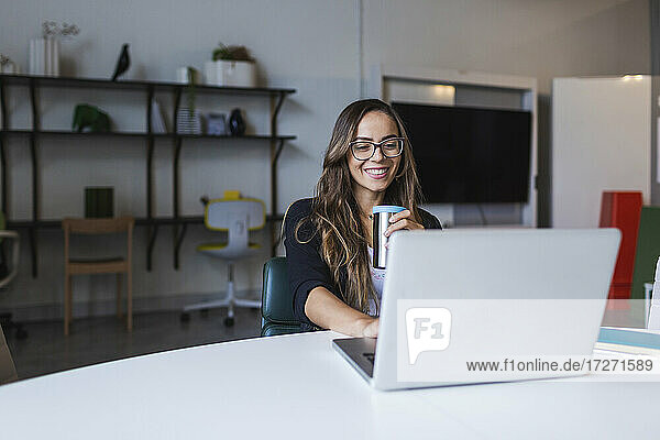 Lächelnde Geschäftsfrau  die Kaffee trinkt und einen Laptop am Schreibtisch im Büro benutzt