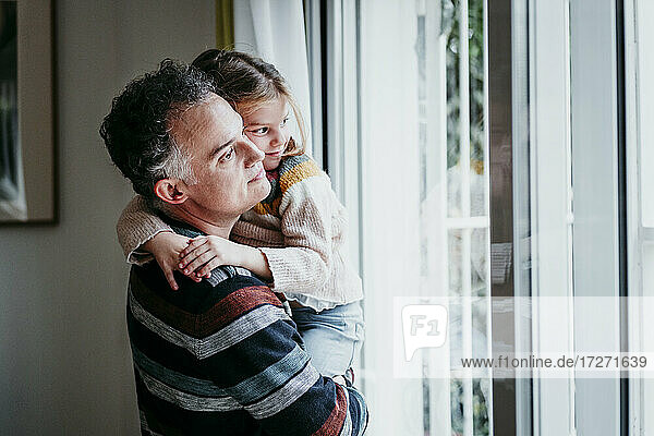 Father carrying daughter while standing by window at home