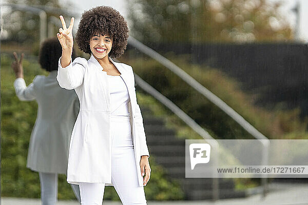Smiling businesswoman showing peace sign while standing against glass wall in city