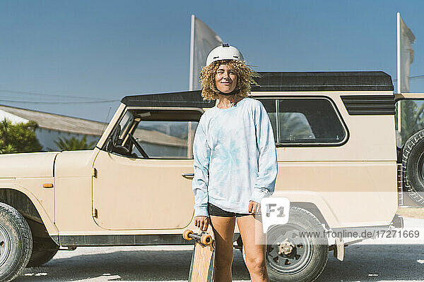 Smiling blond woman wearing helmet holding skateboard while standing against off-road vehicle on sunny day