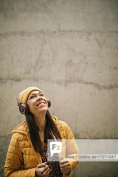 Happy mature woman in warm clothing looking up while listening music through headphones against wall