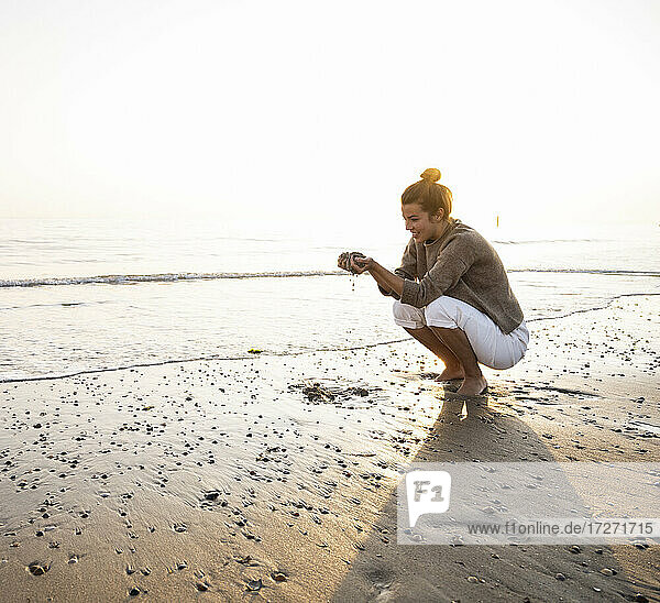 Schöne junge Frau hockt und hält Sand am Strand während des Sonnenuntergangs