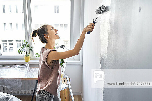 Lächelnde Frau  die zu Hause stehend eine Wand mit einem Farbroller bemalt
