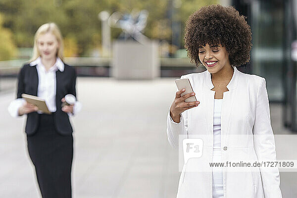 Geschäftsfrau  die ein Mobiltelefon benutzt  während sie mit einem Kollegen im Hintergrund auf dem Fußweg steht