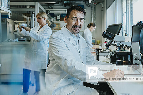 Lächelnder männlicher Wissenschaftler  der am Mikroskop sitzt  während im Hintergrund ein Mitarbeiter im Labor arbeitet